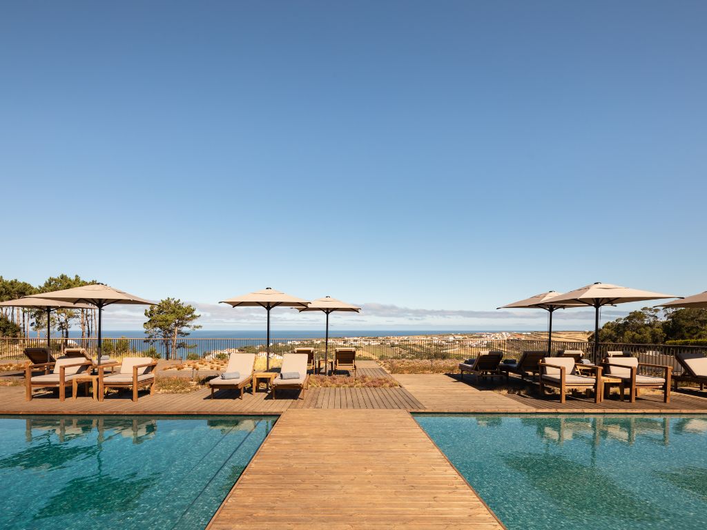 Beach loungers and umbrellas line the pool deck at Immerso Hotel in Ericeira, with the pool in the foreground and the ocean in the background