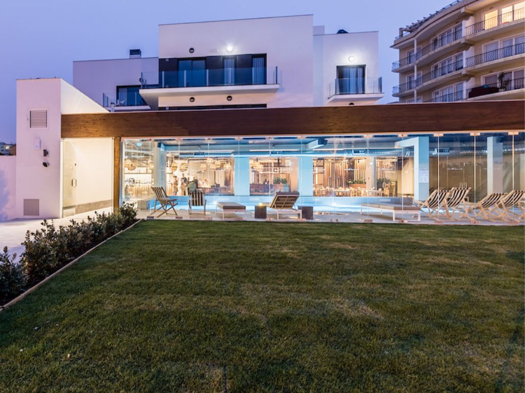 The luxurious pool and garden area of You and The Sea Hotel in Ericeira at sunset. There are striped lounge chairs and the pool has a blue glow