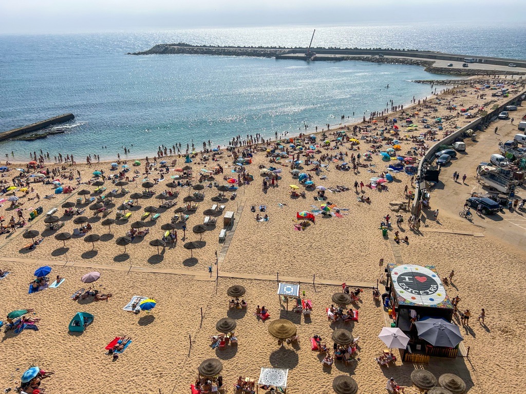 A crowded beach day at Praia dos Pescadores. There are hundreds of families under umbrellas with beach blankets and umbrellas available for rent. There's a beach bar with "I heart Ericeira" painted on the roof