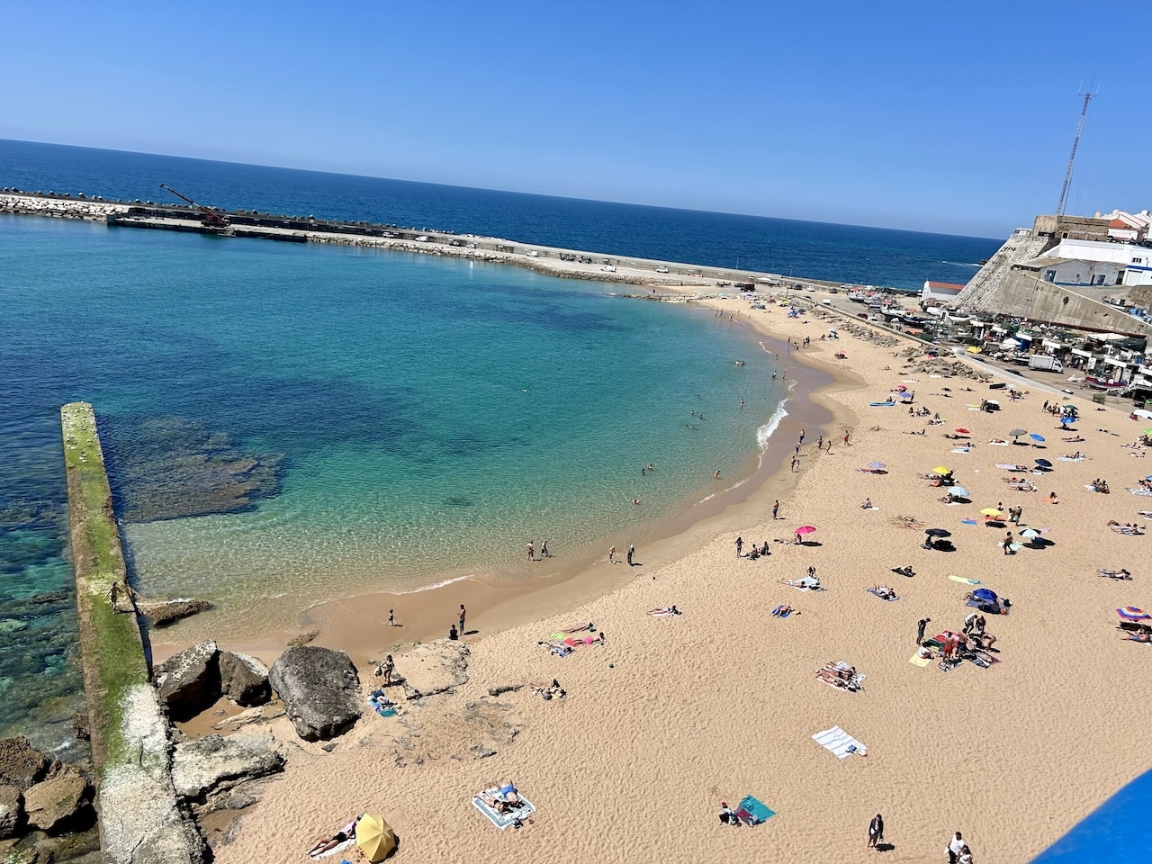 Praia dos pescadores beach on a sunny day in Ericeira. Sunbathers sit on towels and under umbrellas on a c shaped bay